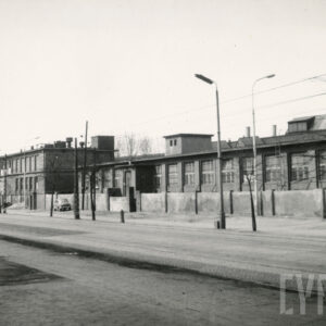 szeroka ulica, wzdłuż niej betonowe ogrodzenie, za nim długie, niskie hale fabryczne