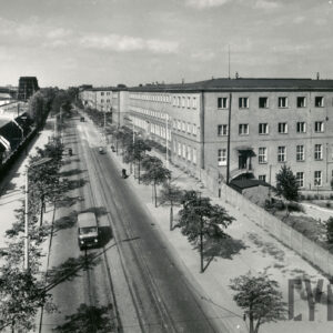 widok z lotu ptaka na brukowaną ulicę z torami tramwajowymi, obsadzoną drzewami, z lewej hale fabryczne, z prawej czterokondygnacyjny (od ulicy o jedną kondygnację niższy) budynek biurowy