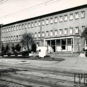 trzypiętrowy budynek z bocznym skrzydłem i wysuniętym przeszklonym wejściem pośrodku bryły, na pierwszym planie ulica z torami tramwajowymi