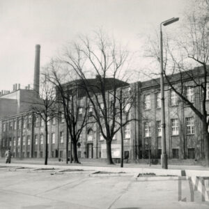 trzykondygnacyjny budynek ceglany, z podwyższonym o jedną kondygnację fragmentem budynku, przed nim drzewa pozbawione liści