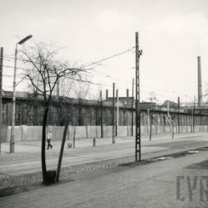 szeroka ulica, wzdłuż niej betonowe ogrodzenie, za nim długie, niskie hale fabryczne