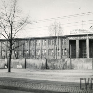 kilkukondygnacyjny budynek ceglany z wysuniętym w partii środkowej wejściem z czterema kolumnami, na górze napis „fabryka obrabiarek", powyżej hasło propagandowe, przed budynkiem betonowe ogrodzenia