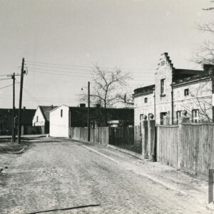 ulica brukowana, przy ulicy z prawej za drewnianym płotem (w części siatka metalowa) jednokondygnacyjny budynek murowany, ceglany z ozdobnym szczytem, w głębi murowane budynki stojące szczytem do ulicy