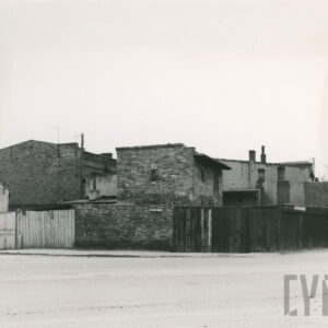 na pierwszym planie asfaltowa ulica, w głębi ogrodzenie w części drewniane, w części murowane, za nim dwu lub jednokondygnacyjna zabudowa murowana, część budynków otynkowana, część ceglana