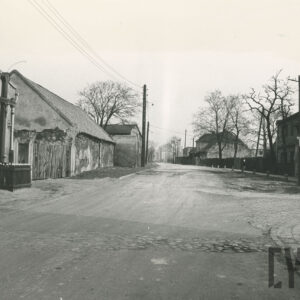 gruntowa ulica, w części widoczny fragmentami bruk, po obu stronach niska zabudowa mieszkalno-gospodarcza, z lewej, przy szczytowej ścianie murowanego budynku drewniany krzyż, ogrodzony drewnianym płotem