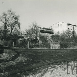 na pierwszym planie gruntowa droga, w głębi ogrodzenie z metalowej siatki, za nim dwa kilkukondygnacyjne budynki mieszkalne, z lewej niższy budynek ceglany, z prawej wyższy murowany, otynkowany, dookoła drzewa i krzewy bez liści