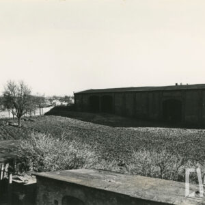 na pierwszym planie fragmenty drewnianej zabudowy gospodarczej, w głębi fragment pola, za nim budynek gospodarczy, z lewej w oddali dom mieszkalny