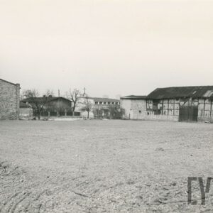 na pierwszym planie fragment ziemi uprawnej zaoranej, w głębi niskie budynki gospodarcze, z lewej murowany, z prawej o konstrukcji szachulcowej