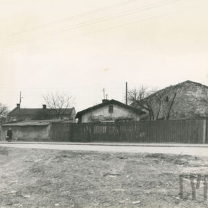 na pierwszym planie niezagospodarowany teren, dalej ulica, w głębi drewniane ogrodzenie, za nim niskie budynki murowane i ceglane