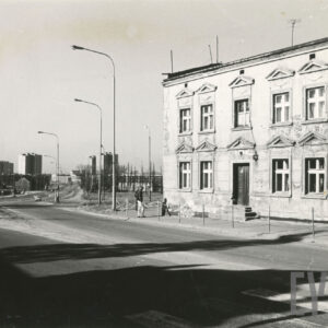 na pierwszym planie asfaltowa ulica, z prawej jednopiętrowy murowany budynek mieszkalny, w głębi kilkunastokondygnacyjne bloki mieszkalne