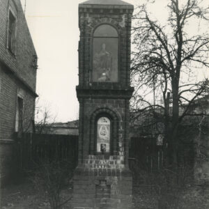 ceglana kapliczka z figurą Jezusa w górnej części i obrazem Matki Boskiej w dolnej zwieńczona krzyżem, po lewej fragment domu, po prawej drzewo