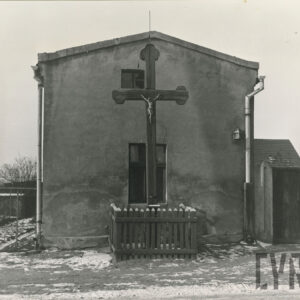 drewniany krzyż z figurą Jezusa ogrodzony drewnianym płotkiem na tle ściany szczytowej domu z dwoma oknami