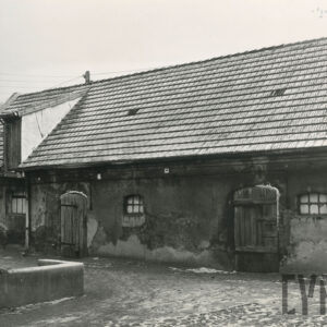 budynek gospodarczy częściowo otynkowany, zniszczony, ze spadzistym dachem pokrytym dachówką, małe okienka, drewniane drzwi