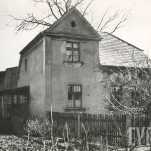 otynkowany dom w ogrodzie z dachem częściowo przykrytym dachówką, częściowo zwieńczony szczytem, z lewej strony drewniana przybudówka, na pierwszym planie drewniany płot