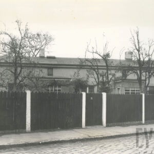 fragment brukowanej ulicy, za nią drewniany płot, za płotem murowany dom ze spadzistym dachem