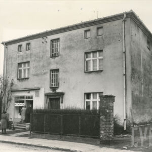 piętrowy, murowany dom z poddaszem i sklepem spożywczym w parterze, przed nim fragment drucianego płotu obrośniętego krzewami