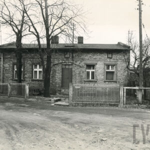 ceglany, parterowy budynek ze spadzistym dachem, drzwiami na osi i oknami po bokach częściowo ogrodzony płotem z metalowych prętów