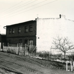 bita, rozjechana ulica, wzdłuż niej druciany płot, za płotem ceglany budynek z fasadą z oknami i ślepym szczytem