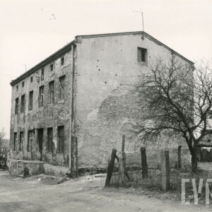 murowany, pokryty odpadającym tynkiem piętrowy dom z poddaszem i spadzistym dachem, po prawej fragment ogrodu otoczonego drucianym płotem