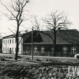 rozkopana ulica, w głębi dwa domy mieszkalne, po lewej piętrowy z rzędami okien, po prawej parterowy kryty spadzistym dachem