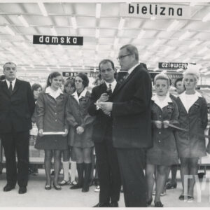 wnętrze dużego sklepu, na pierwszym planie przemawia mężczyzna, obok niego stoi mężczyzna z mikrofonem, za nimi kobiety w fartuszkach z białymi kołnierzykami i kilku mężczyzn w garniturach