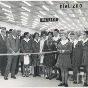 wnętrze dużego sklepu, duża grupa kobiet i mężczyzn, część z kobiet ubrana w fartuszkach z białymi kołnierzykami, pośrodku wstęga przygotowana do przecięcia