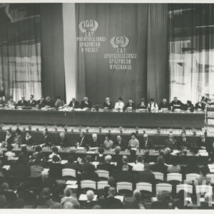 w głębi długi stół na podwyższeniu, przy nim siedzi kilkanaście osób, poniżej, poniżej drugi stół przy nim siedzi kilkanaście osób, na widowni, tyłem do zdjęcia siedzą w rzędach ludzie