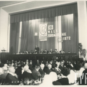 w głębi długi stół na podwyższeniu, przy nim siedzi kilka osób, jedna przemawia w pozycji stojącej, poniżej długie stoły, stojące prostopadle do tego na podwyższeniu, przy nich siedzą ludzie