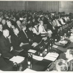 w długich rzędach siedzą ludzie, na przeciwko nich długi stół przy którym siedzi kilkanaście osób zwróconych do pozostałych twarzami