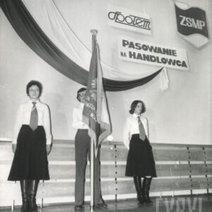 mężczyzna stoi ze sztandarem, dwie kobiety po bokach, za nimi udrapowana tkanina (w kolorach polskiej flagi)