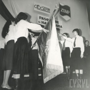 grupa dziewcząt w strojach galowych stoi po obu stronach sztandaru z wyciągniętą do przysięgi prawą ręką