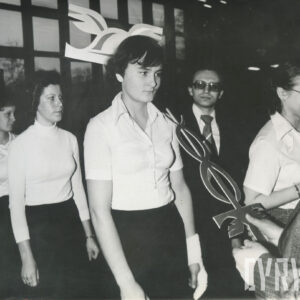 trzy młode kobiety w galowych strojach (jasne bluzki, ciemne spódnice), pierwsza z nich w trakcie pasowania