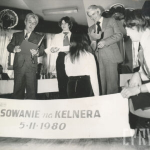 dwóch mężczyzn, w środku młoda kobieta, tyłem klęczy dziewczyna, na pierwszym planie baner o treści „pasowanie na kelnera, 5.11.1980”