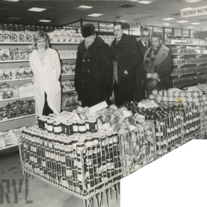 wnętrze sklepu, pośrodku kosze ze słoikami i puszkami, za nimi z lewej kobieta w białym fartuchu, obok niej czterech mężczyzn w płaszczach, w głębi półki z artykułami