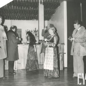 grupa ludzi stoi przed barem w lokalu, dwie kobiety (kelnerki) w długich sukniach, mężczyźni (klienci) w płaszczach