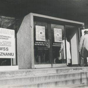 oszklone wejście do budynku, z lewej tablica informacyjna, przez otwarte drzwi wchodzą dwie osoby