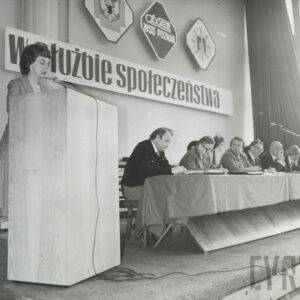 na pierwszym planie kobieta przemawia na mównicy, za nią długi stół przy którym siedzi kilka osób, za nimi na ścianie powieszona dekoracja i napis „w służbie społeczeństwa”