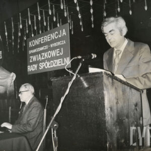 mężczyzny przemawia na mównicy, z lewej mężczyzna siedzi przy stole, za nimi tablica z nazwą konferencji