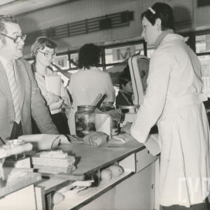 z lewej mężczyzna i kobieta przy ladzie, za ladą z prawej kobieta w jasnym fartuchu, na lodzie towary