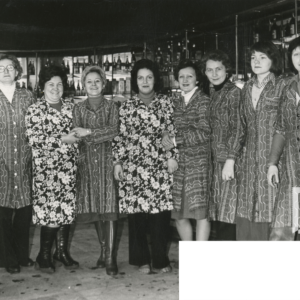 dziewięć kobiet we wzorzystych fartuchach stoi pośrodku sklepu i pozuje do zdjęcia, za nimi półki z artykułami