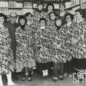 wnętrze sklepu, dziesięć kobiet w bardzo wzorzystych fartuch, między nimi jeden mężczyzna, za nimi półki z artykułami