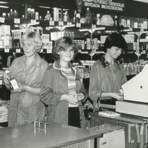 trzy kobiety w fartuchach, za ladą, za nimi półki z artykułami drogeryjnymi