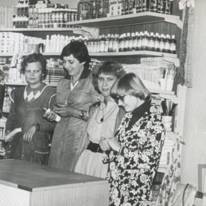 cztery kobiety w fartuchach stoją za ladą sklepową, za nimi półki z artykułami