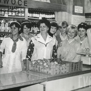 siedem kobiet w fartuchach stoją za ladą sklepową, za nimi półki z artykułami, na ladzie waga i plastikowe pojemniki z owocami i warzywami