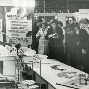 kobieta w stroju pielęgniarki stoi przy stoliku, na którym leży lalka (niemowlę), z prawej kobiety i mężczyźni przyglądają się pokazowi