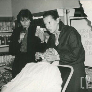 na fotelu leży kobieta, nakryta białą peleryną, dwie kobiety przy jej głowie, jedna z nich wykonuje zabieg kosmetyczny