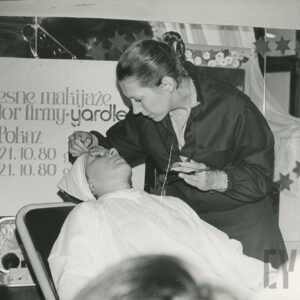 na fotelu leży kobieta, nakryta białą peleryną, nad nią stoi kobieta, która wykonuje jej makijaz