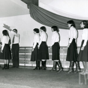 grupa kobiet w jasnych bluzkach i ciemnych spódnicach stoi w jednym szeregu, podchodząc do kobiety i mężczyzny