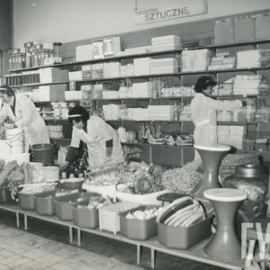 wnętrze sklepu, na pierwszym planie niski stolik, na nim wystawione artykuły na sprzedaż, za nim trzy kobiety w białych fartucha układają towary na półkach, za nimi, w głębi półki z artykułami na sprzedaż