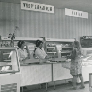 wnętrze sklepu, na pierwszym, w jednym szeregu lady z dwiema wagami i lady chłodnicze, przy nich dwie ekspedientki w białych fartuchach, przy ladzie klientka z koszykiem, w głębi półki z towarem